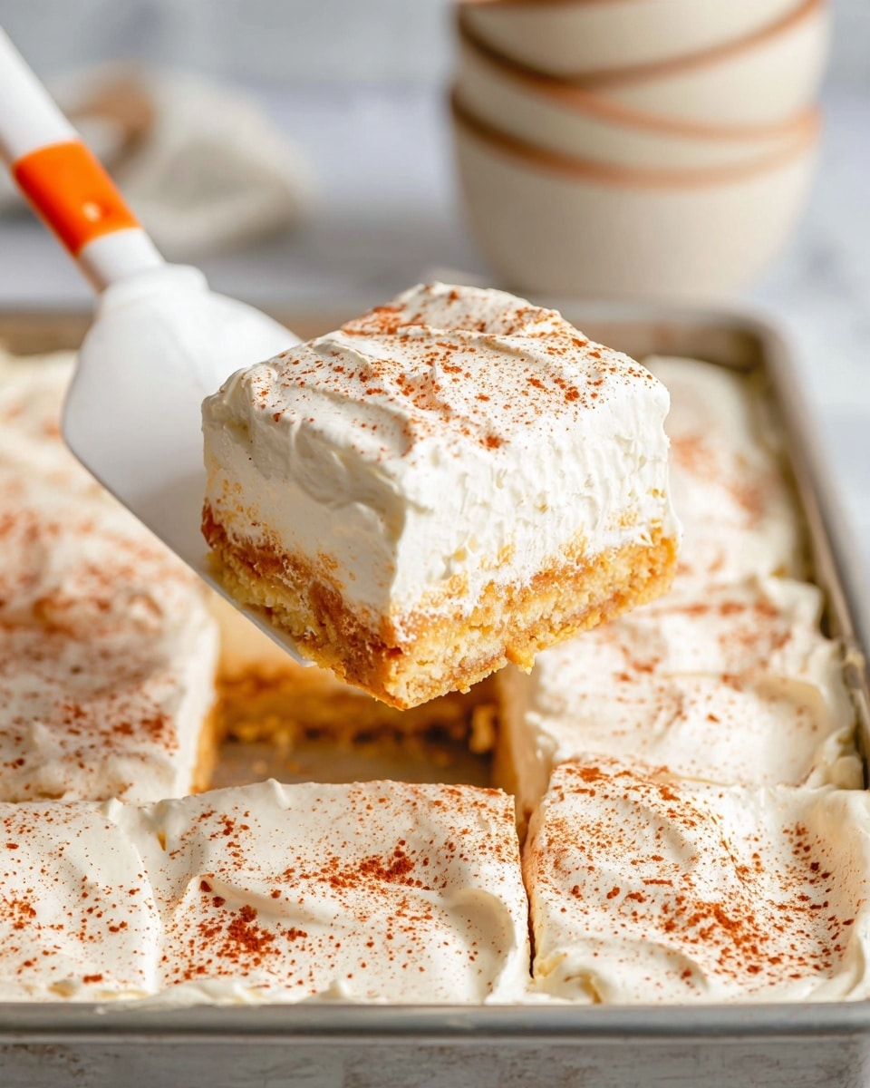 A close-up of a square dessert piece being lifted from a tray with a white spatula that has an orange tip. The dessert has two visible layers; the bottom layer is golden-brown and textured, and the top layer is thick white whipped cream sprinkled evenly with fine reddish-brown powder. The tray holding many squares of the dessert is metal, with other pieces showing the same two layers and powder topping. The background is a white marbled surface with blurred pale bowls stacked behind. photo taken with an iphone --ar 4:5 --v 7