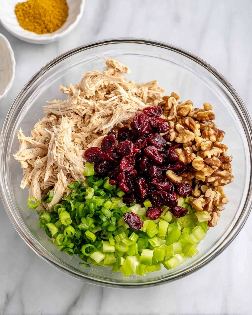 A clear glass bowl shows six main layers of ingredients arranged side by side. On the left is a layer of light beige shredded chicken with soft texture. Next to it are bright green chopped scallions, fresh and finely sliced. In the center, a pile of dark red dried cranberries with a slightly wrinkled texture fills the middle. To the right of the cranberries is a layer of light brown chopped walnuts with a rough texture. On the far right is a layer of pale green diced celery, cut into small even cubes. The bowl sits on a white marbled surface, and in the top left corner outside the bowl, a small white dish holds a bright golden-yellow powder. Photo taken with an iphone --ar 4:5 --v 7