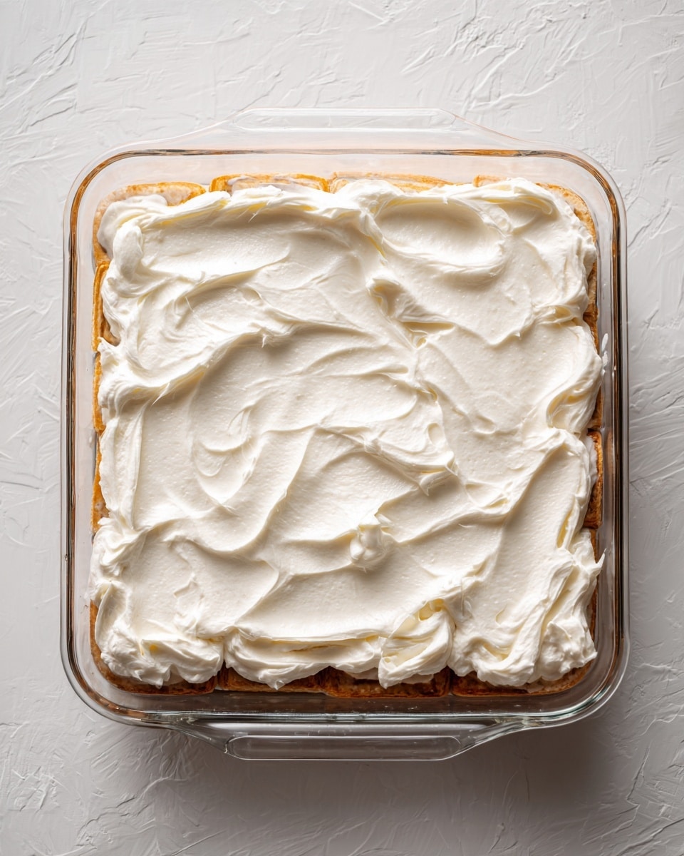 A clear glass rectangular baking dish holds small rolled pastries with a marbled light brown and cream pattern arranged along the edges. In the center, there is a thick, creamy white layer of frosting spread smoothly but with visible swirls and slight texture. The dish sits on a white marbled surface, and the lighting shows a shiny, fresh look on the frosting. photo taken with an iphone --ar 4:5 --v 7