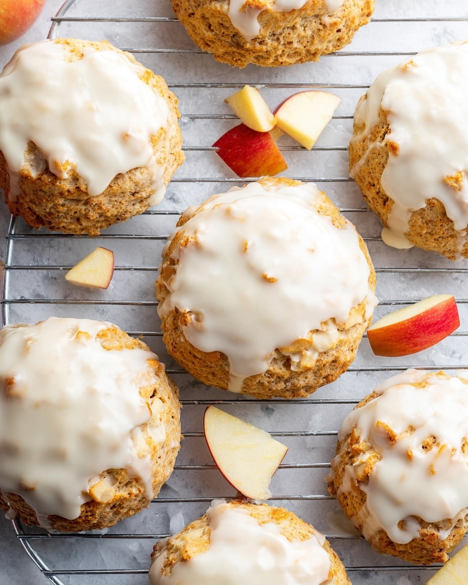 The image shows several round scones with a light golden-brown textured base and a thick white icing layer on top that looks smooth and slightly drippy, covering most of each scone’s surface. The scones are placed on a silver wire cooling rack over a white marbled texture background. To the side, there are thin slices and small cubes of red-skinned apples adding a fresh touch to the scene. photo taken with an iphone --ar 4:5 --v 7