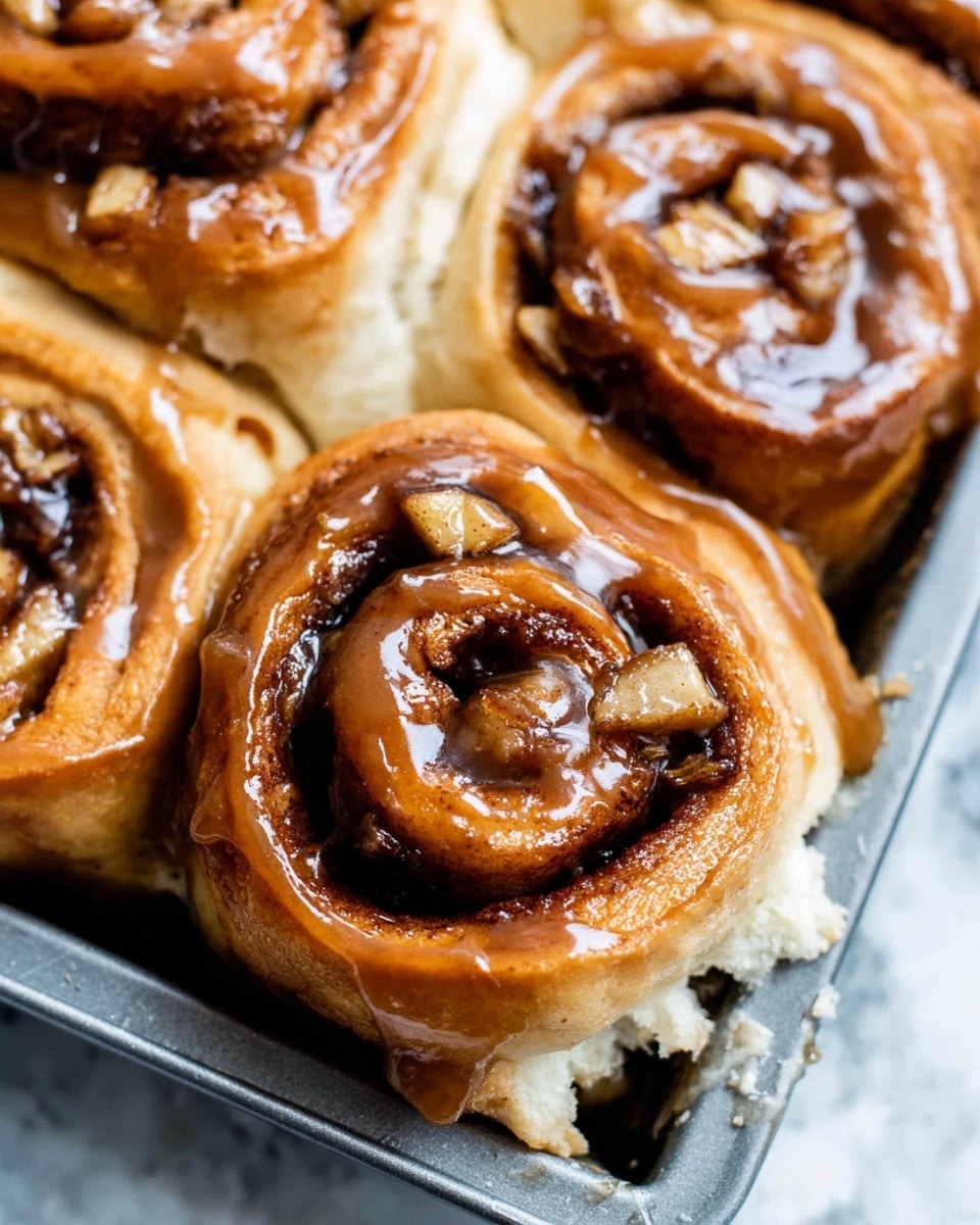 The image shows a tray full of unbaked cinnamon rolls arranged close together, each roll having three or four visible layers of soft dough spiraled tightly. Between the dough layers, there is a dark brown cinnamon filling with a slightly glossy texture. Small chunks of green apple and bits of chopped nuts are scattered unevenly inside the rolls, peeking out from the swirls. The rolls are pale and smooth, with a soft dough texture, sitting in a metal pan on a white marbled surface. The close-up perspective highlights the detail of each roll’s layers and fillings, photo taken with an iphone --ar 4:5 --v 7