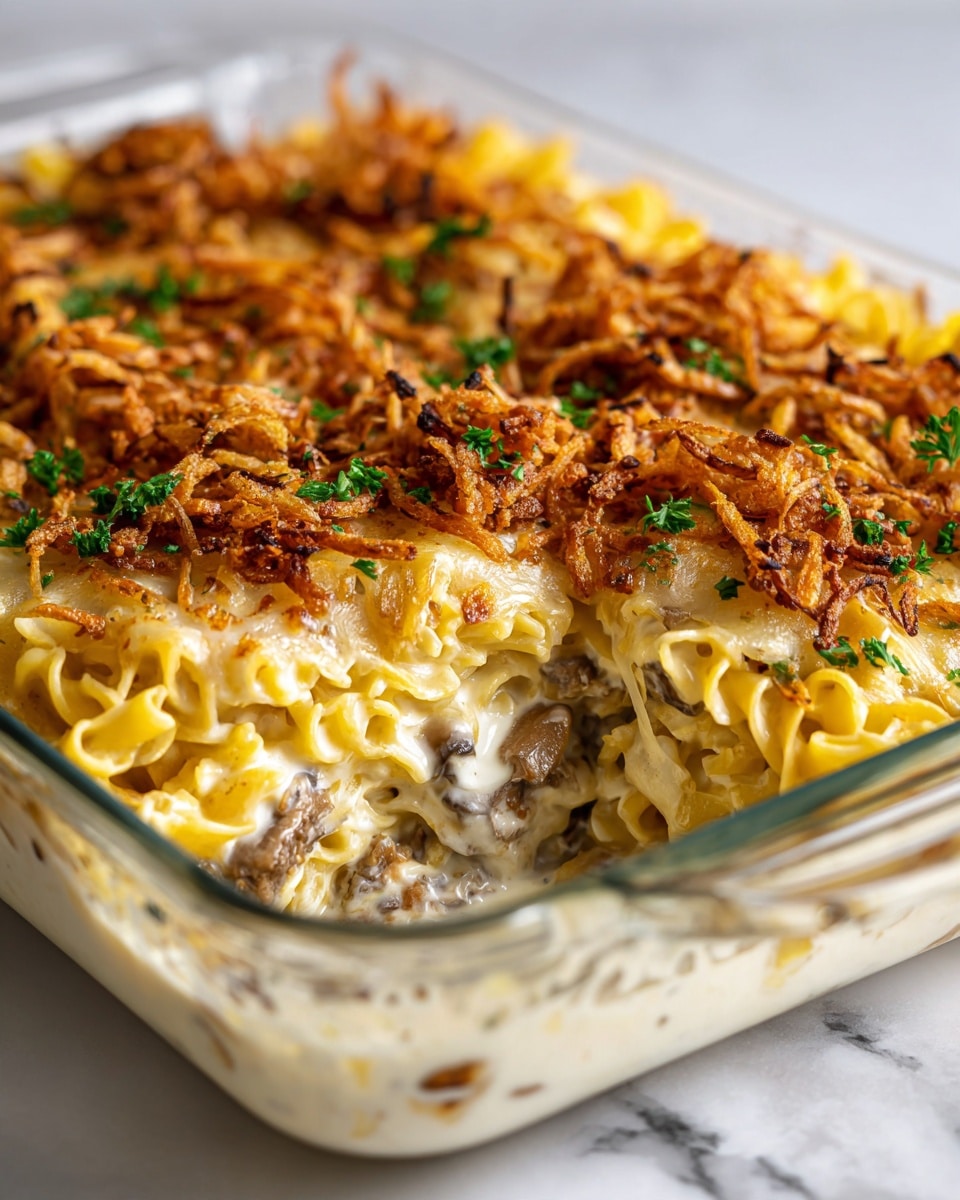 A close-up image showing a baked casserole in a clear rectangular dish on a white marbled surface. The dish has three visible layers: a creamy white sauce mixed with mushrooms and noodles at the bottom, yellow egg noodles forming a thick middle layer, and a top layer of golden brown crispy fried onions scattered all over. Small green parsley pieces are sprinkled on top, adding a touch of color. A silver serving spoon is partially inside the dish, lifting some of the creamy noodles from the corner. Photo taken with an iphone --ar 4:5 --v 7