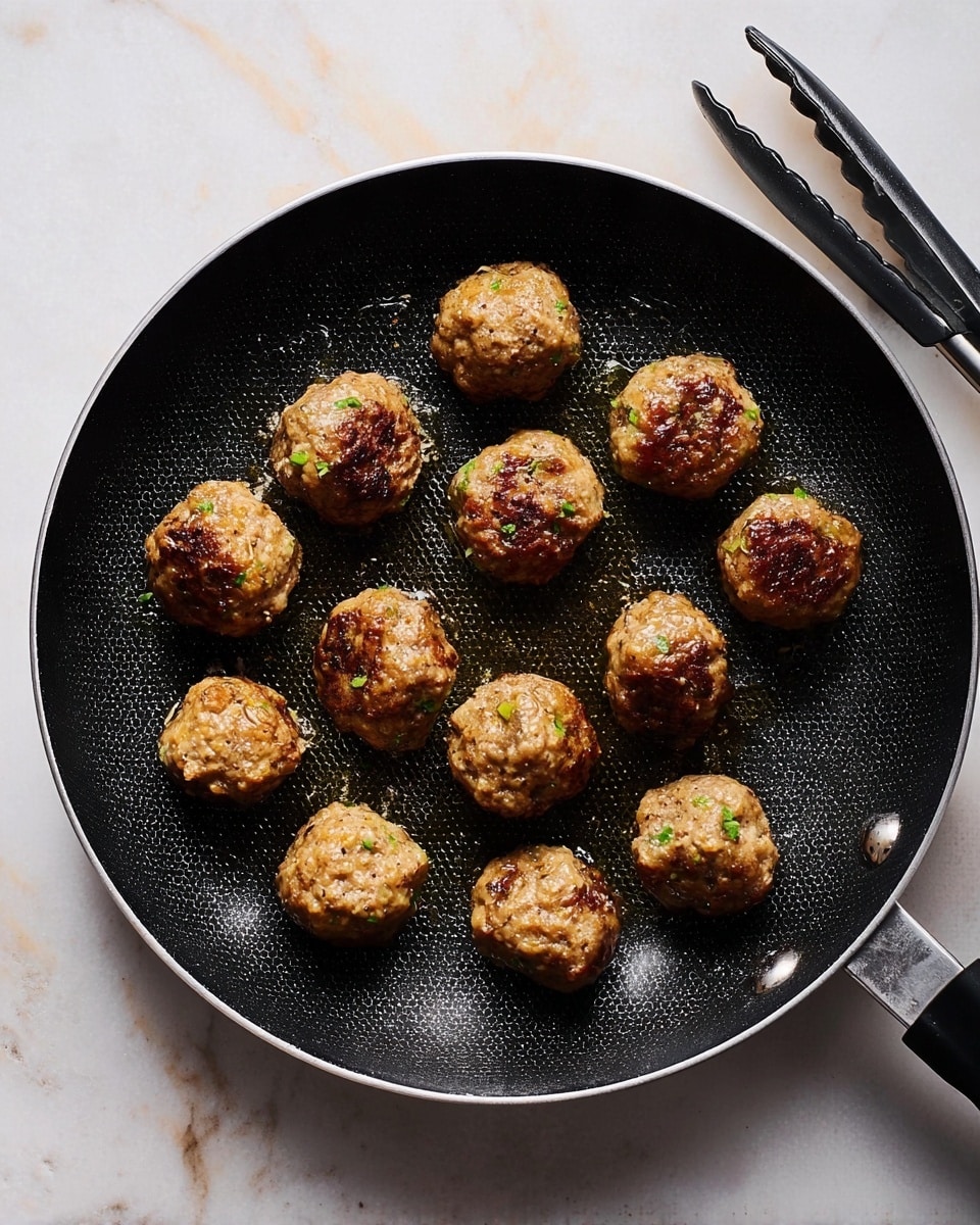A black frying pan with a silver handle holds thirteen golden-brown meatballs, each with a slightly crispy texture and small green herb flecks visible on their surface. The meatballs are arranged unevenly across the pan's honeycomb pattern coating, and a small amount of oil glistens around them. The pan rests on a white marbled surface, with a pair of black tongs placed nearby on the right side. photo taken with an iphone --ar 4:5 --v 7