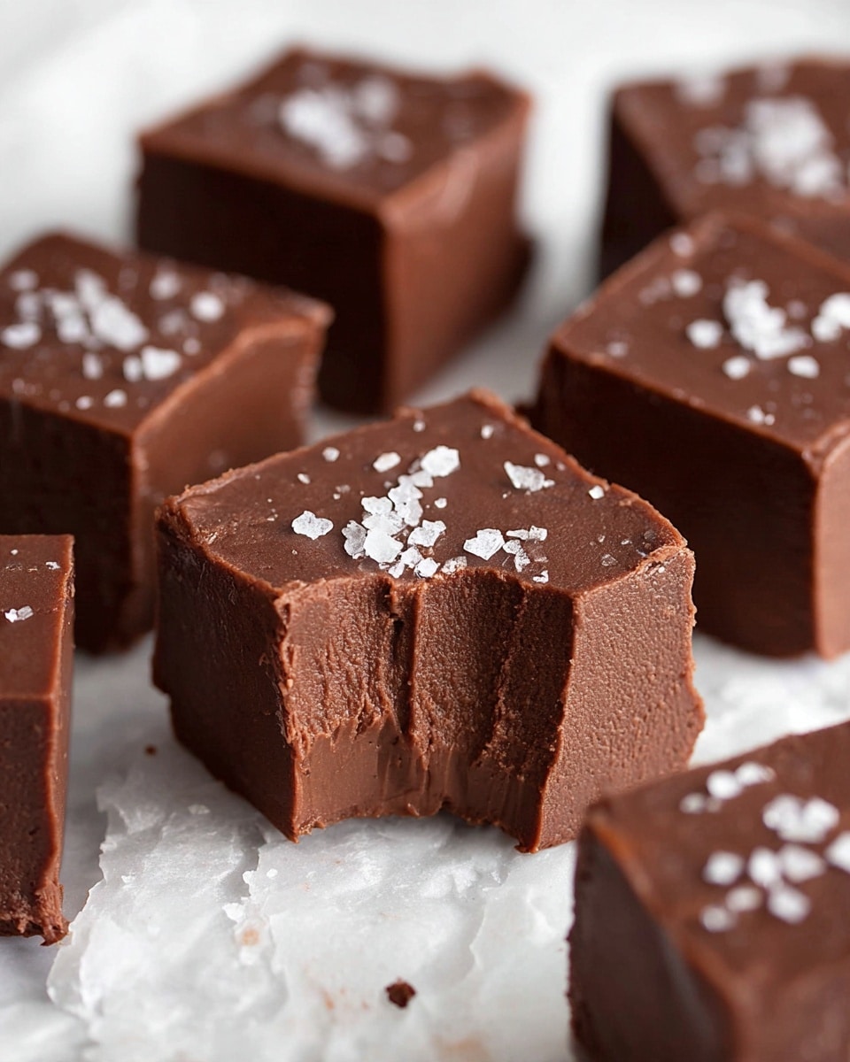 The image shows several thick square pieces of rich dark brown chocolate fudge on a white marbled texture surface. Each piece has a smooth and slightly shiny top with small crystals of coarse salt sprinkled on it. One piece in the front has a bite taken out, showing a dense, creamy and slightly lighter brown inside layer with a soft texture. The fudge pieces are arranged irregularly with some blurred in the background, highlighting the sharp focus on the bitten piece in front. Photo taken with an iphone --ar 4:5 --v 7