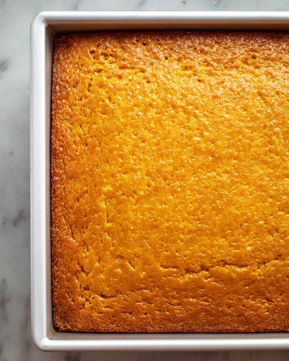 A close-up top view of a single-layer orange-brown baked cake with a slightly rough texture on its surface, sitting inside a white rimmed rectangular baking pan. The edges of the cake show slight browning, indicating a crisp border, while the middle remains evenly cooked with a warm golden hue. The pan is placed on a white marbled surface that provides a clean, light background. photo taken with an iphone --ar 4:5 --v 7