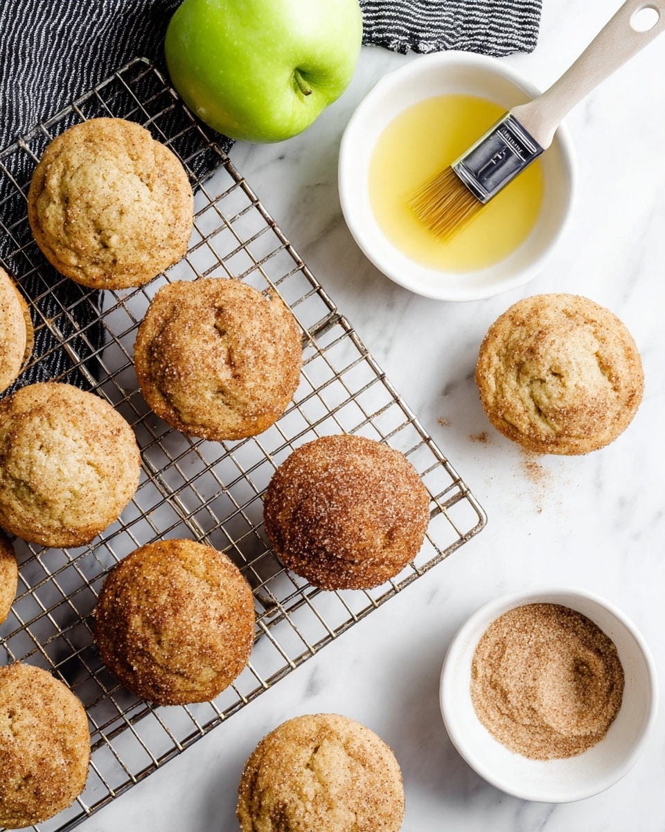 A wire cooling rack sitting on a white marbled surface holds ten light brown muffins with slightly cracked tops, arranged scattered on and off the rack. Two muffins near the center are coated with a slightly darker brown sugar and cinnamon topping, creating a textured crust. To the top right, a small white bowl filled with yellow melted butter holds a brush resting inside. Next to this is another small white bowl filled with a cinnamon and sugar mix, smooth and powdery in texture. A bright green apple lies on the top left corner, resting on a black and white striped cloth. Photo taken with an iphone --ar 4:5 --v 7