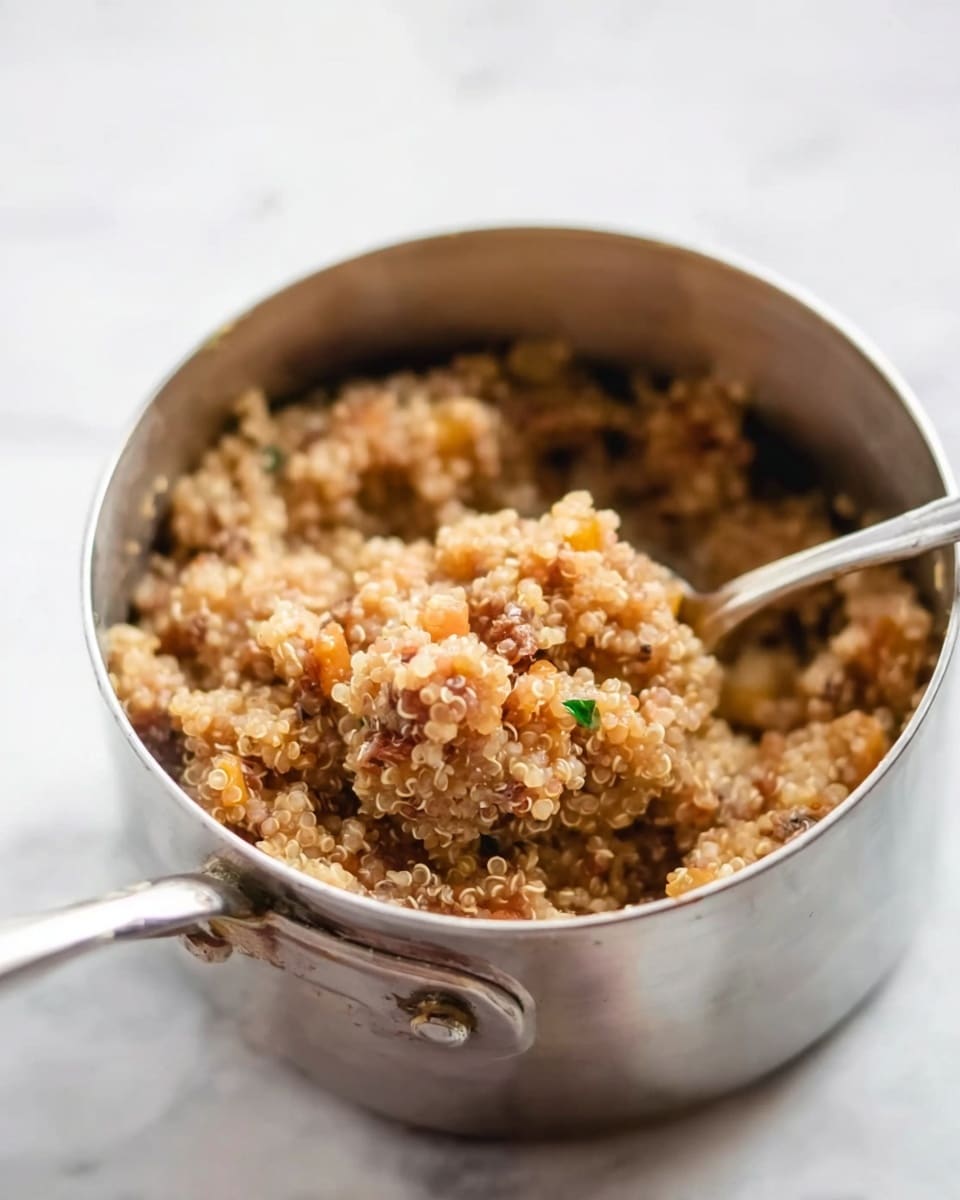A close-up of a small silver pot filled with cooked quinoa mixed with small chunks of soft light brown vegetables, showing a fluffy and slightly grainy texture. Inside the pot, there is a silver spoon resting, partially covered with the quinoa mixture. The pot is placed on a white marbled surface, and steam rises gently from the mixture, suggesting it is warm. photo taken with an iphone --ar 4:5 --v 7