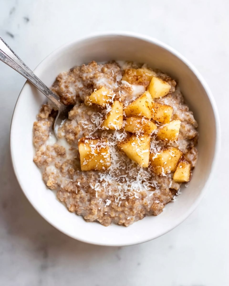 A white bowl filled with a soft, thick porridge that has a light brown, grainy texture covering the bottom and middle layers. On top, there are small, irregular chunks of light golden-brown fruit pieces sprinkled with fine white shreds, likely coconut. A silver spoon is partially buried in the porridge on the left side of the bowl. The bowl sits on a white marbled surface. photo taken with an iphone --ar 4:5 --v 7