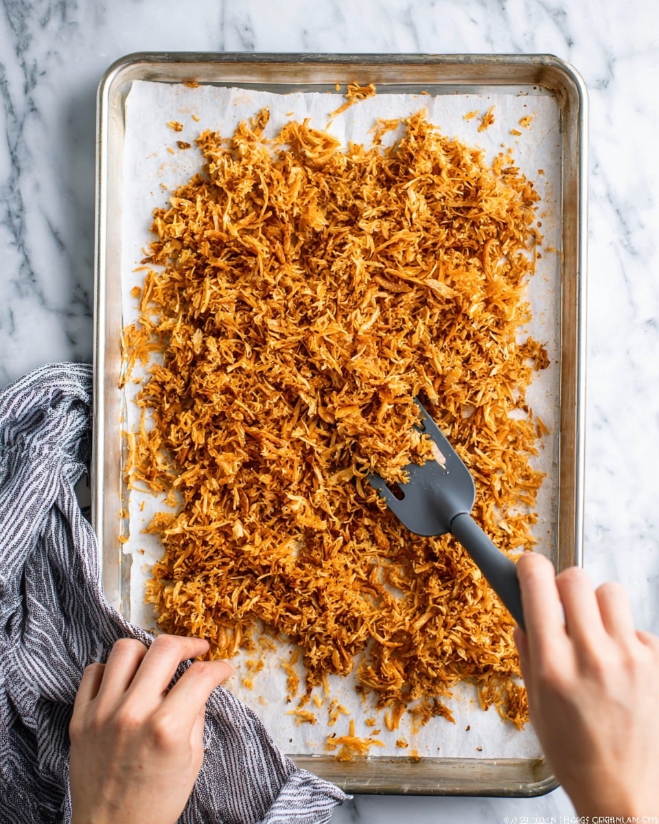 A metal baking tray lined with white parchment paper holds a spread of thin, shredded, golden-brown cooked pieces with a slightly crispy texture. The pieces are loosely scattered over most of the tray, with some smaller bits around the edges. A woman's hand holding a gray spatula with black handle is gently moving the shredded pieces, showing the uneven clumps and different shades of brown and orange from seasoning. Another woman's hand is visible holding a striped gray cloth near the bottom left corner of the tray. The tray sits on a white marbled surface. photo taken with an iphone --ar 4:5 --v 7