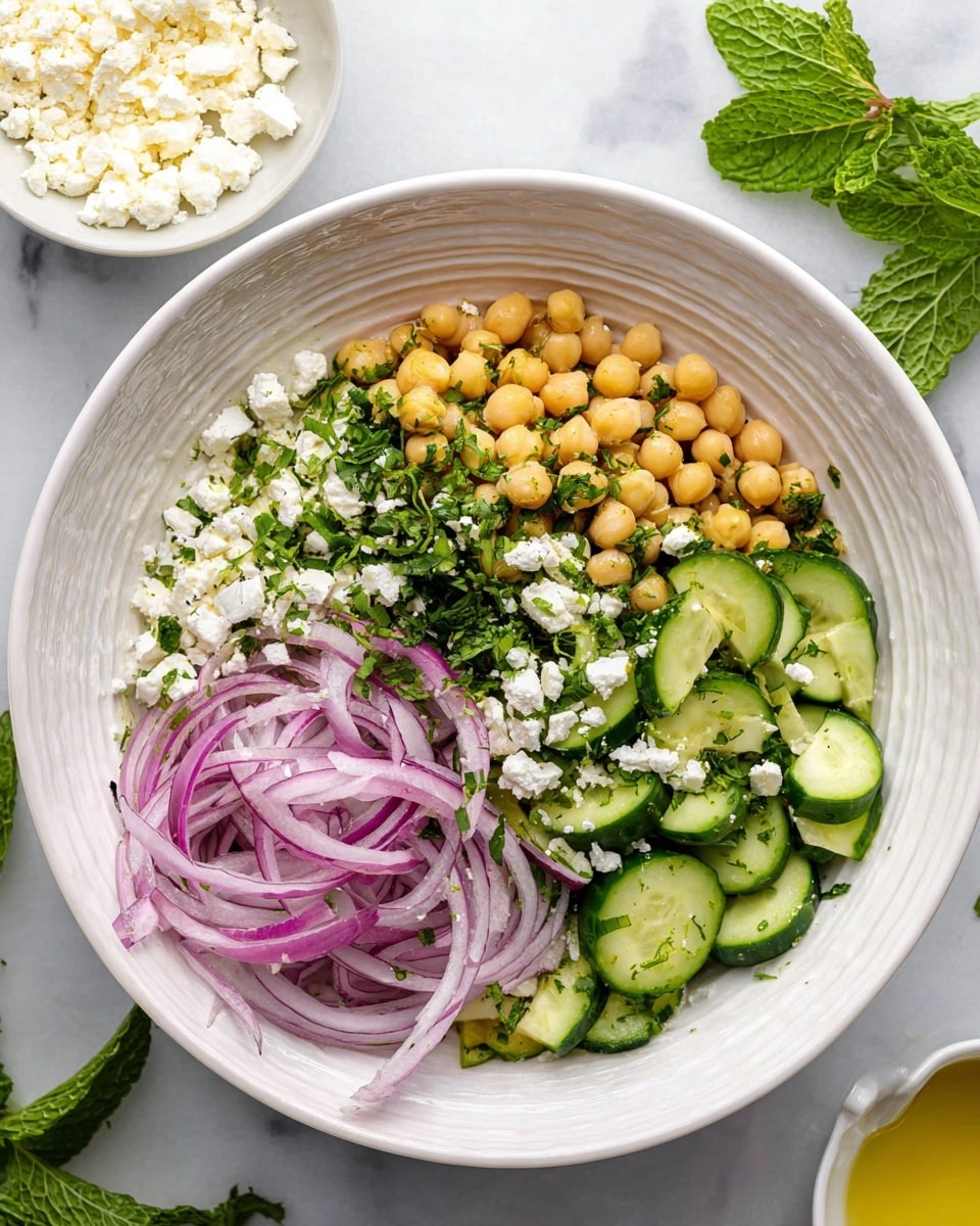 A white bowl with ridged sides holds a salad with four main sections: pale yellow chickpeas forming a chunky layer on the top right, thin sliced purple onions layered diagonally in the middle, finely chopped green herbs sprinkled both over the onions and the bottom left, and fresh sliced cucumber pieces forming the bottom left section. Crumbled white cheese is scattered on top mostly over the onions and chickpeas. The bowl sits on a white marbled surface with fresh green mint leaves beside it. A small white bowl with more crumbled cheese is near the top left corner and a white dish with yellow dressing is at the bottom right corner of the frame. Photo taken with an iphone --ar 4:5 --v 7
